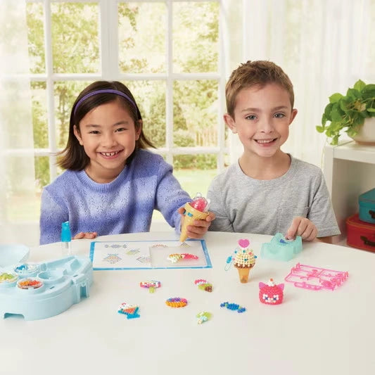 Two children making colourful Aquabeads ice cream creations at a table, with the ice cream bead pen, bead trays, sprayer and finished designs displayed in front of them.