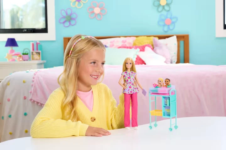 Smiling blonde girl in a yellow cardigan sitting at a white table with the Barbie Baby Doctor doll and turquoise examination trolley holding two baby dolls, in a bedroom with pink bedding.