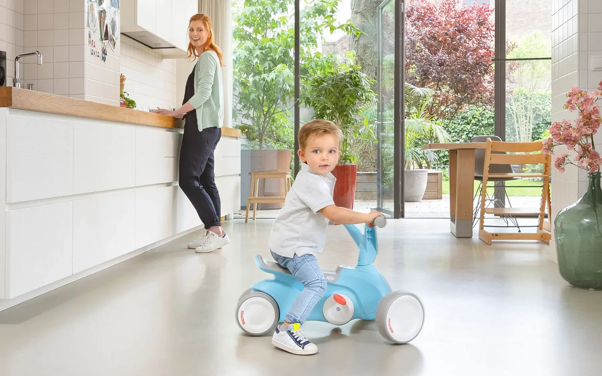 Child seated on a blue BERG GO² ride-on indoors with an adult standing near a counter