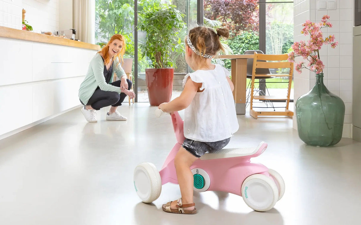 Child seated on pink BERG GO² ride-on indoors with adult crouching nearby