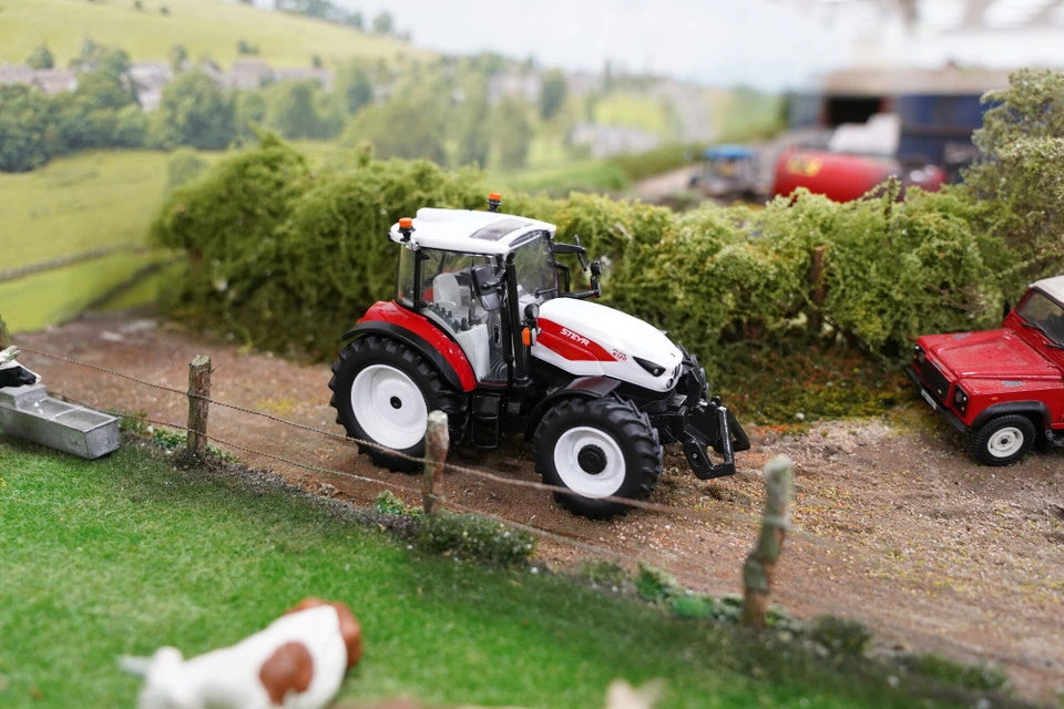 Britains Steyr 4120 Plus farm toy tractor on a model farm lane with a red trailer and cows visible