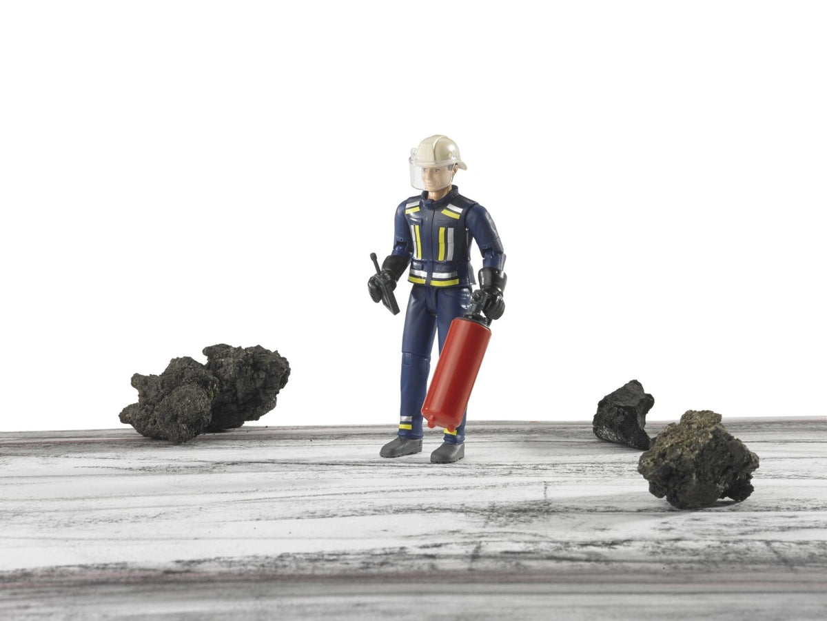 Bruder bworld Fireman with Accessories 60100 standing on a wooden surface among dark rocks, holding a red fire extinguisher and black radio while wearing protective firefighter clothing