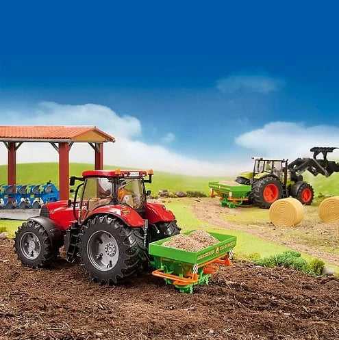 A red toy tractor in a farm scene pulling a green Amazone fertiliser spreader filled with material.