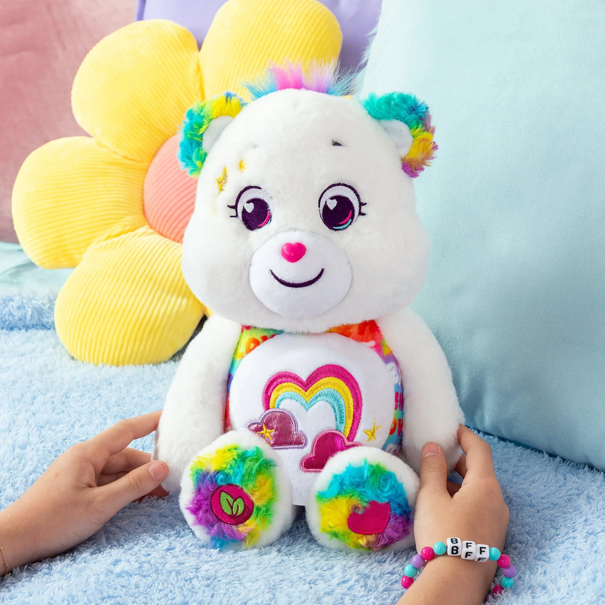 Care Bears True Friend Bear plush on a bed with colourful ears and paws surrounded by soft pillows in Ireland.