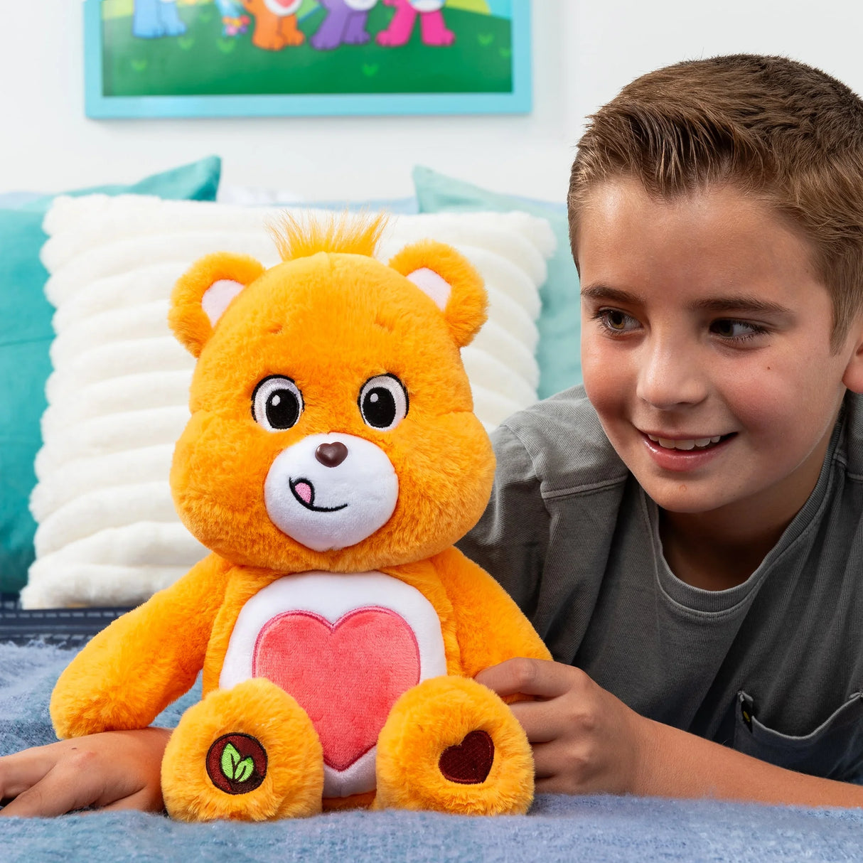 Child sitting on bed holding Care Bears Tenderheart Bear plush with bright orange fur and red heart belly badge in Ireland.
