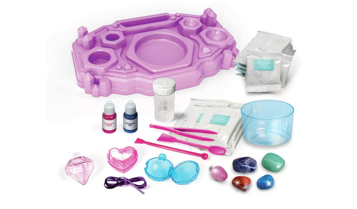Purple Crystal Jewellery Creator workstation surrounded by chemical packets, blue and pink colour bottles, measuring cups, pink tweezers, spoon, spatula, ribbon, gemstone specimens and jewellery charms.