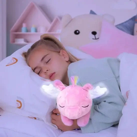 Young girl sleeping in bed holding the pink Poppy Pegasus plush, showing its closed embroidered eyes and illuminated white wings.