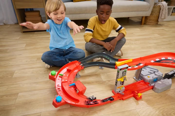 Two boys sitting on a wooden floor playing with the Hot Wheels Formula 1 Downhill Circuit Race set, featuring the red and black curved racing track, three miniature F1 cars and upright lap counter.