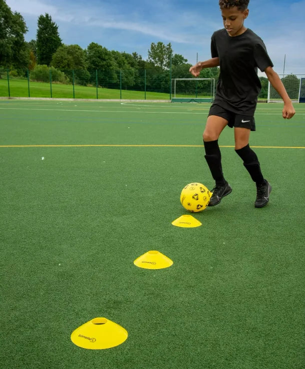 Child on an outdoor artificial pitch controlling a yellow football between three yellow training cones