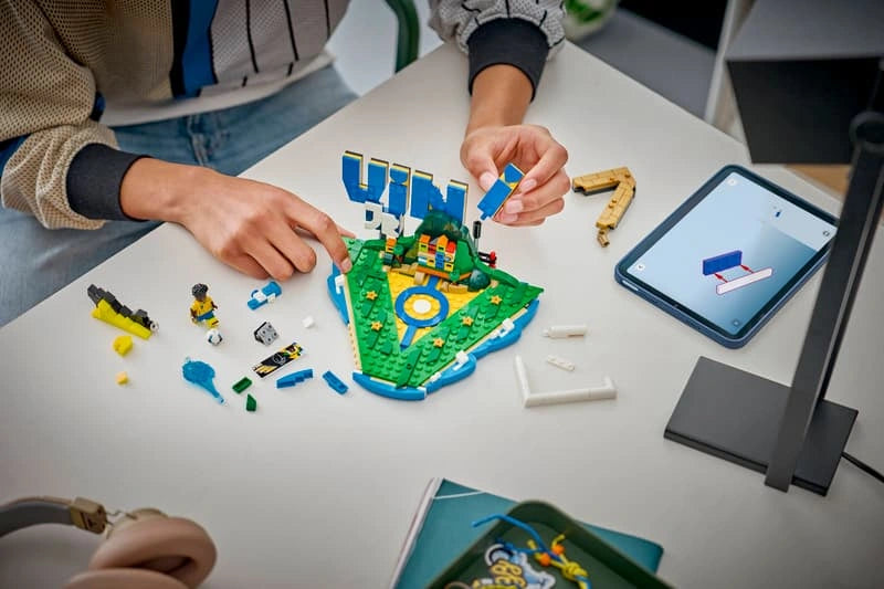 Person assembling LEGO Editions Vini Jr. Football Highlights 43027 on a table with loose LEGO bricks, a partially completed football display and tablet showing digital building instructions.