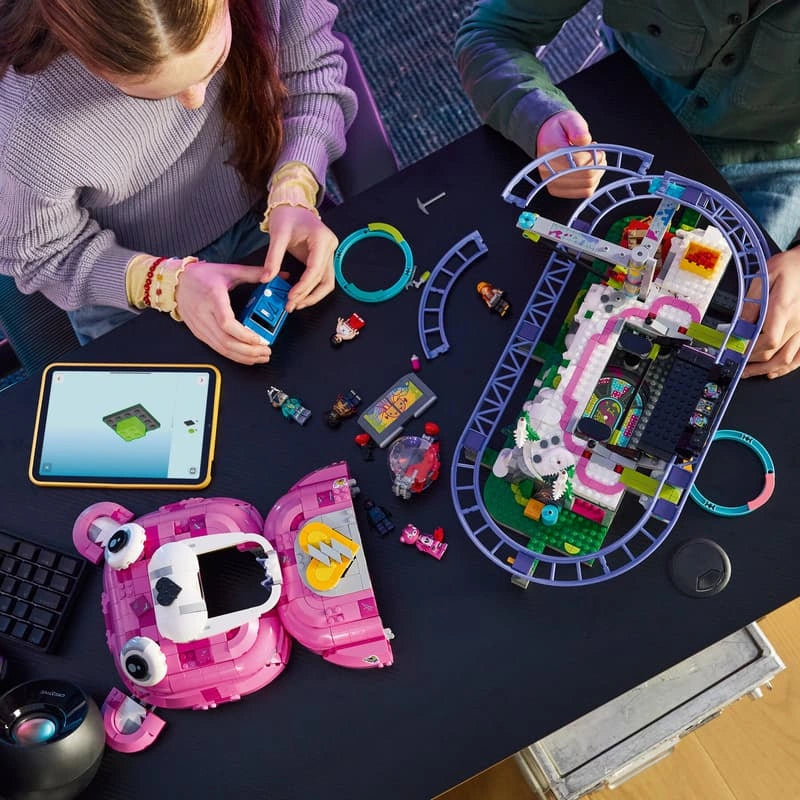 Overhead view of two children assembling LEGO Fortnite Rave Cave 77082 with a partially built roller coaster, pink bear head sections, loose bricks and tablet instructions.