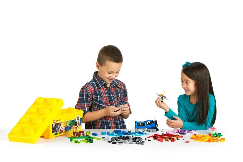 Two children building models with LEGO Medium Creative Brick Box 10696, surrounded by colourful bricks, a blue train and yellow storage box.