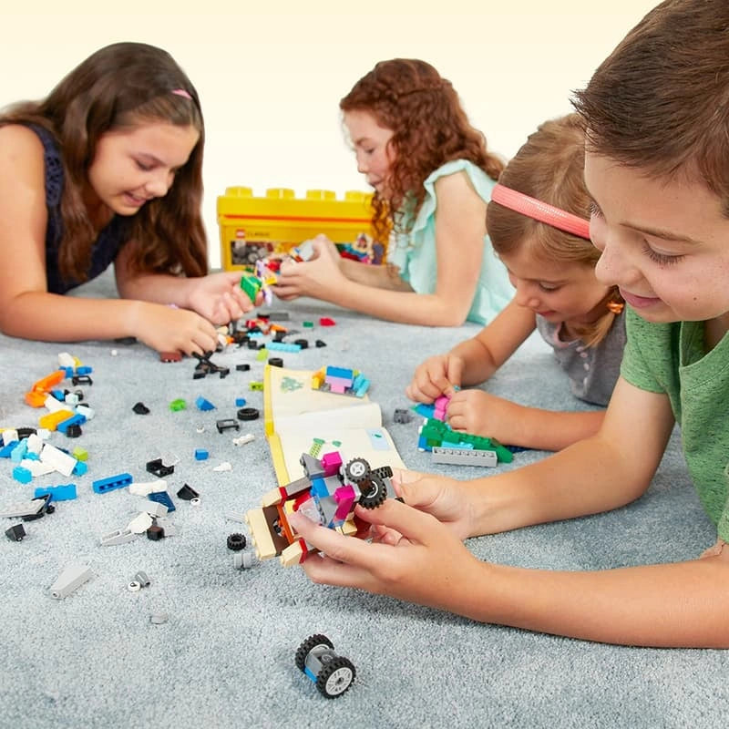 Four children building with LEGO Medium Creative Brick Box 10696, with colourful bricks, wheels and models spread across a carpet.