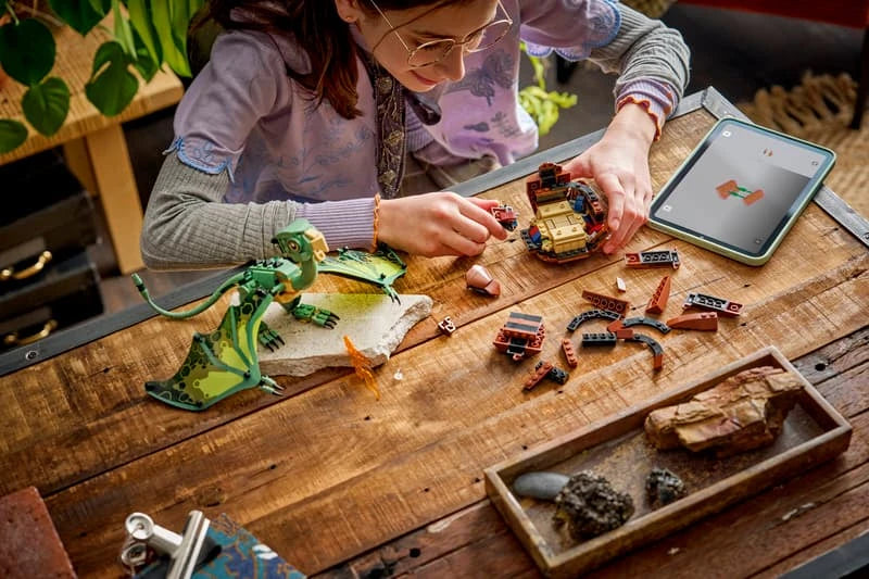 Child building LEGO pieces at a table with a tablet and green dragon model nearby