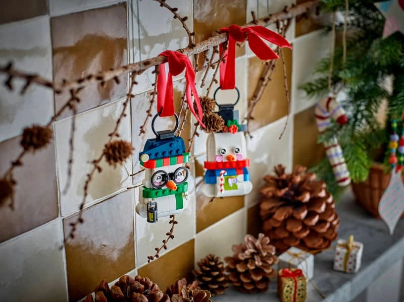 Two LEGO snowman ornaments hanging by black loops from a twig with red ribbons, pine cones and Christmas decorations