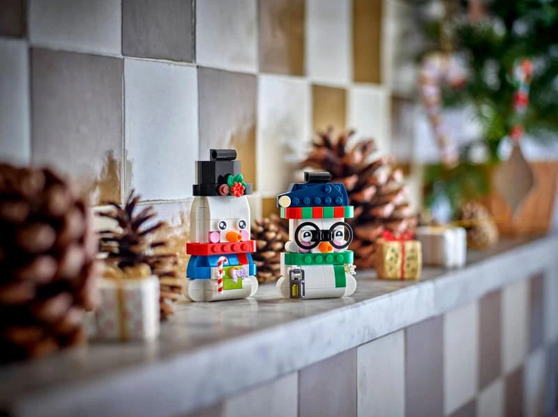Two LEGO snowman ornaments standing on a tiled shelf with pine cones, wrapped gifts and Christmas greenery behind