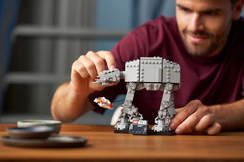 Close-up of a man adjusting the head of the LEGO Star Wars AT-AT model on a wooden table, showing the grey walker, orange and white Snowspeeder and display stand.