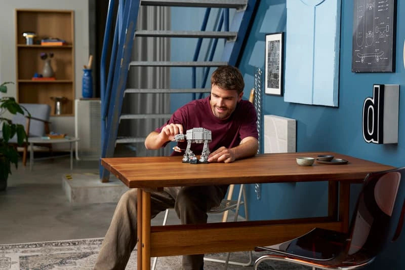 Man adjusting the completed LEGO Star Wars AT-AT walker on a wooden table, with the grey model and black display stand positioned in front of him.