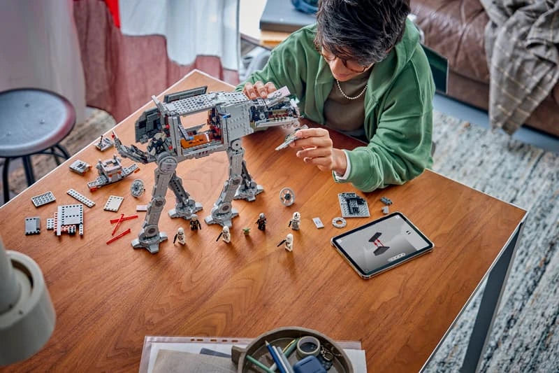 Child building the LEGO Star Wars Imperial Remnant AT-AT with INT-4 set 75454, with loose LEGO pieces, minifigures and a tablet on a wooden table.