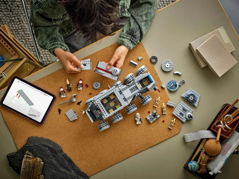 Person assembling the LEGO Republic Juggernaut at a table with loose wheels, vehicle sections, figures and digital instructions