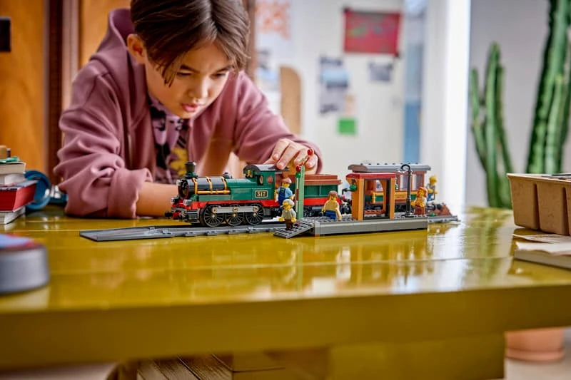 Child building a LEGO City steam train set at a table with train, platform, track pieces and tablet instructions