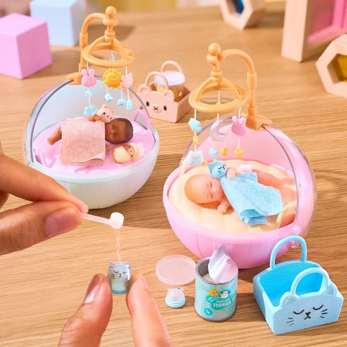 Two Miniverse Real Baby Nurture miniature babies resting in pale blue and pink bassinets with hanging mobiles, while hands use a tiny spoon above a miniature bottle, with a formula tin and blue animal face basket in the foreground.