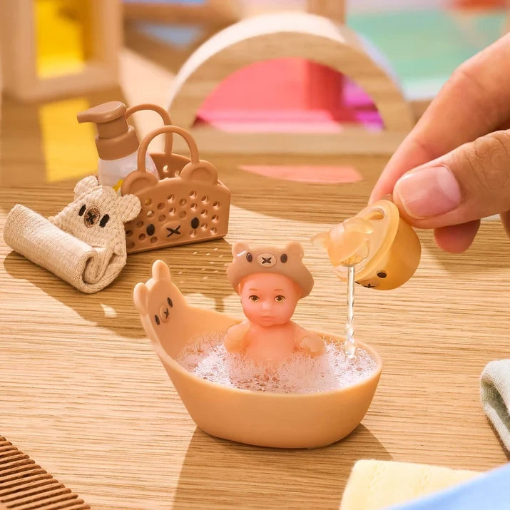 Miniverse Real Baby Nurture miniature baby wearing a brown bear hood and sitting in a tan animal shaped bath filled with foam, while a hand pours water from a matching scoop. A folded towel, bottle and bear face basket sit nearby.