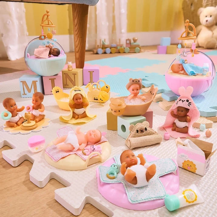 Collection of Miniverse Real Baby Nurture miniature babies arranged on a playroom floor, featuring babies in bassinets with hanging mobiles, a foam filled bath, bunny hooded towels, changing mats, stacking ring toys and miniature baby care accessories.