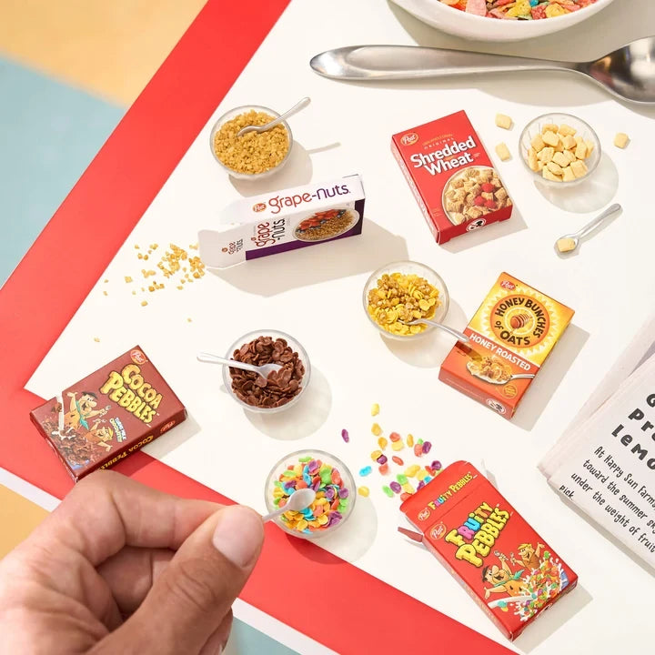 Overhead view of Miniverse miniature cereal boxes and bowls containing Fruity Pebbles, Cocoa Pebbles, Grape-Nuts, Shredded Wheat and Honey Bunches of Oats, with a hand holding a miniature spoon.