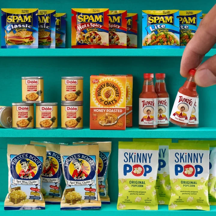 Miniature SPAM packets, Dole pineapple cans, Honey Bunches of Oats cereal, Tapatío hot sauce, Pirate's Booty snacks and SkinnyPop popcorn arranged on turquoise shelves, with a hand lifting a tiny hot sauce bottle.