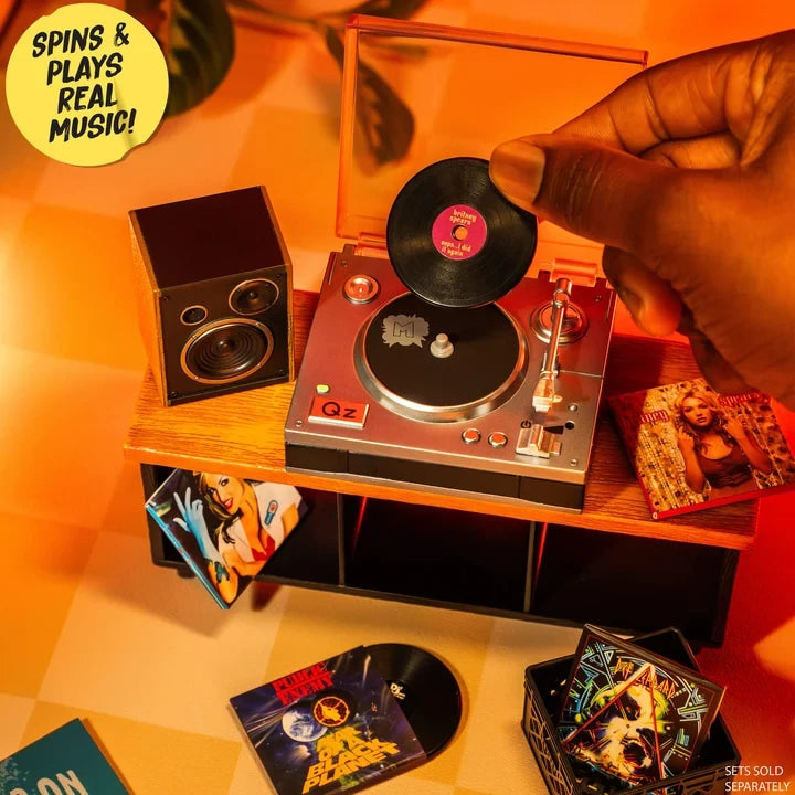 Close-up of a hand placing a miniature black vinyl record onto the Miniverse Real Music Record Player, with miniature speakers, album sleeves and record crates. Record player sold separately.