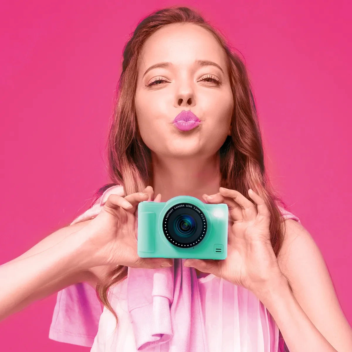 Woman holding the green Photo Creator Pop Colour camera at chest height against a bright pink background, wearing a pink top and posing with pursed lips.