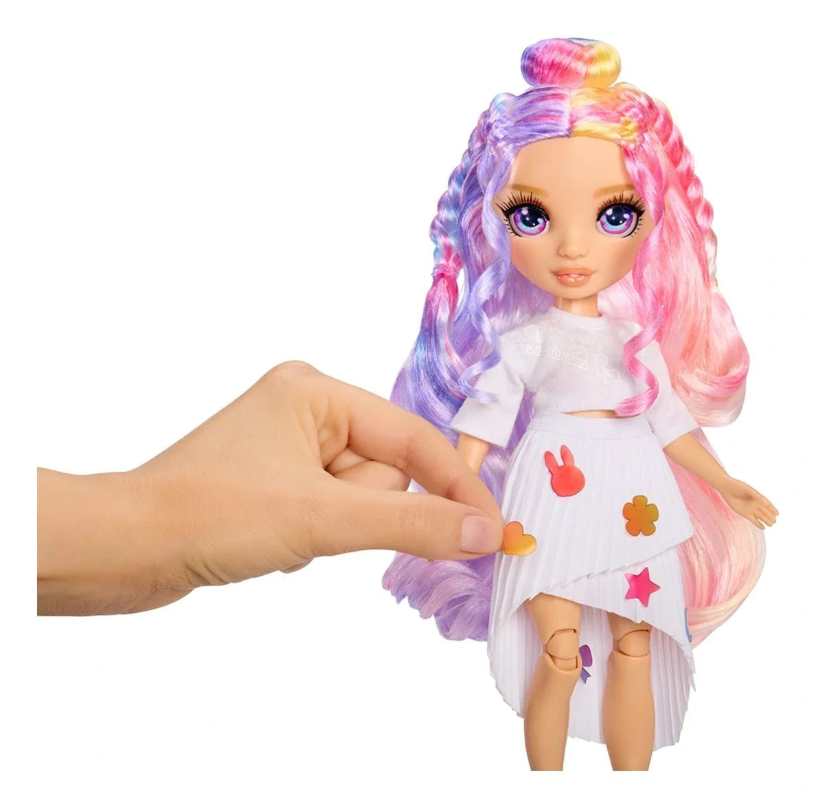 Rainbow High Air Brush & Create doll with pink and purple hair and a white outfit decorated with colourful bunny, flower, heart and star shapes, while a hand applies an orange heart to her skirt.