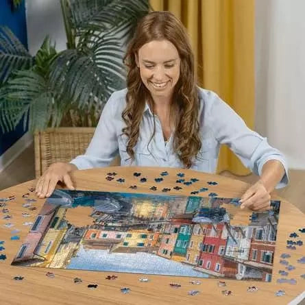 Woman assembling the Ravensburger Burano Italy jigsaw puzzle on a table with loose pieces around the canal scene