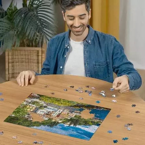 Man assembling the Ravensburger Lake Como Italy jigsaw puzzle on a table with loose pieces around the lakeside scene