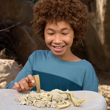 Child using wooden digging tool on table with sand and dinosaur bone pieces, rocky background behind