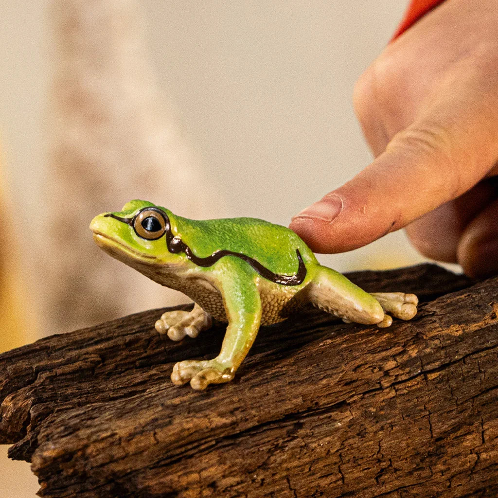 A green tree frog figurine on a piece of wood, shown with a hand beside it for scale and its side stripe clearly visible.