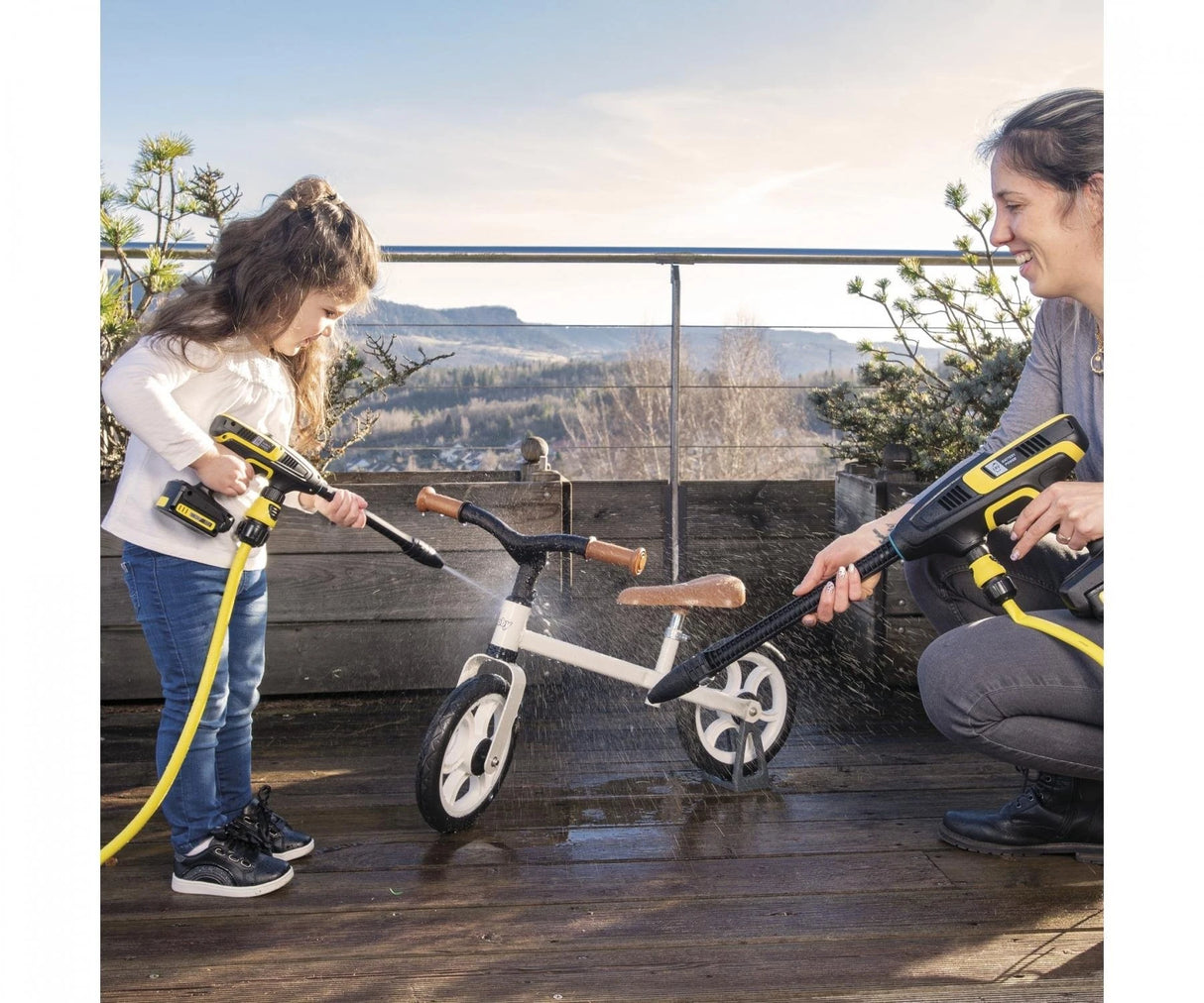 Child and adult using Smoby Karcher High Pressure Gun toys outdoors to clean a balance bike on a decked terrace