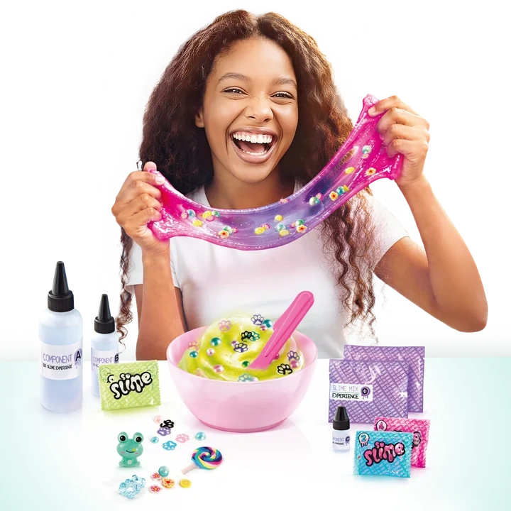 Smiling girl stretching pink and purple slime filled with colourful decorations, with a pink mixing bowl, ingredient bottles, spatula, slime sachets and small charms displayed on the table.