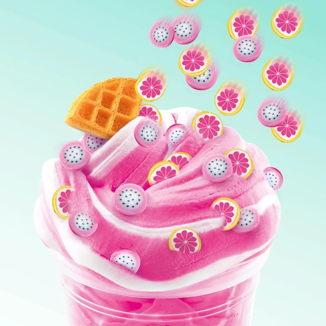 Close-up of pink and white swirled So Slime in a clear pot, decorated with miniature dragon fruit and pink grapefruit slices and a waffle shaped charm against a pale turquoise background.