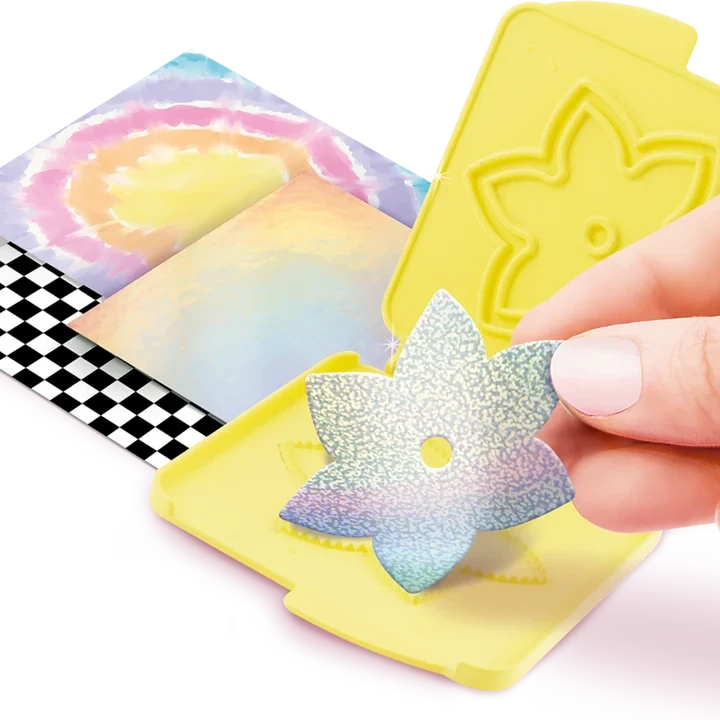 Hand holding an iridescent paper flower cutout above an open yellow flower cutting plate, with pastel coloured paper sheets and black and white checked paper underneath.