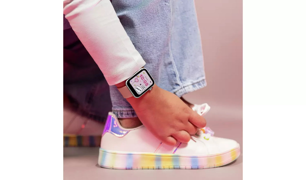 Close-up of a smartwatch with pink digital watch face worn on a wrist beside white trainers with rainbow coloured soles and blue jeans.