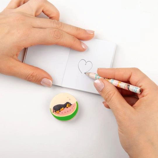Hands drawing a heart in a small notebook with a TOPModel pencil and round illustrated accessory