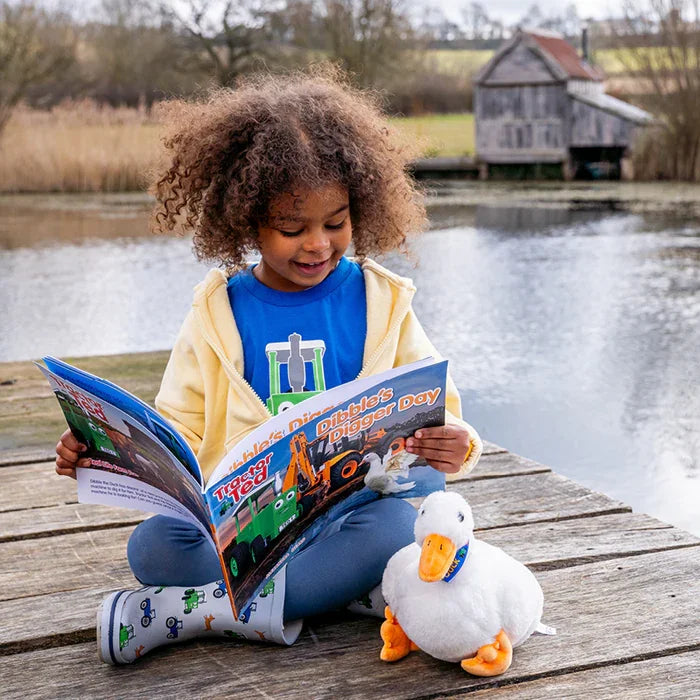 Child sitting by a pond reading the Tractor Ted Dibbles Digger Day storybook with a soft toy duck