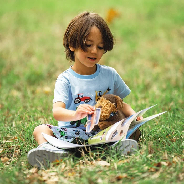 Child sitting on grass with an open Tractor Ted farm activity book, holding a tractor sticker and a soft toy while looking through the illustrated pages.