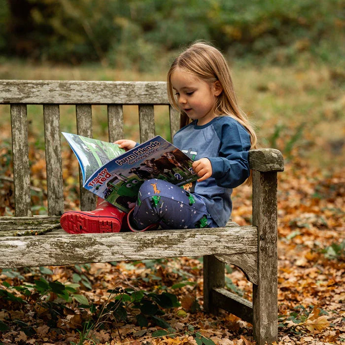 Child sitting outdoors on grass reading Tractor Ted Farm Friends Counting Storybook with farm illustration visible on open pages