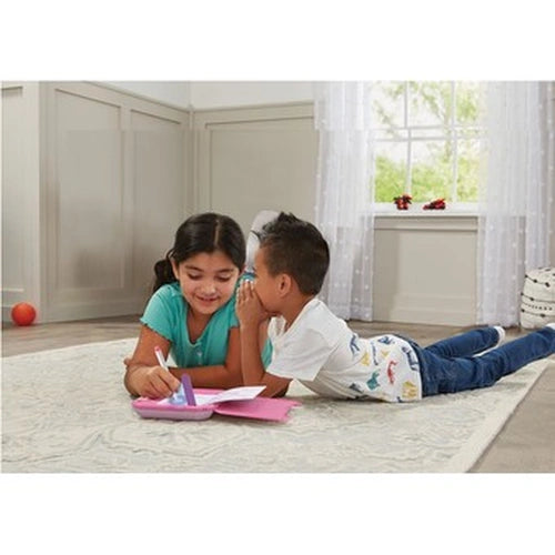 Two children lying on a rug with the VTech Secret Safe Magic Notebook open, one writing while the other whispers in her ear