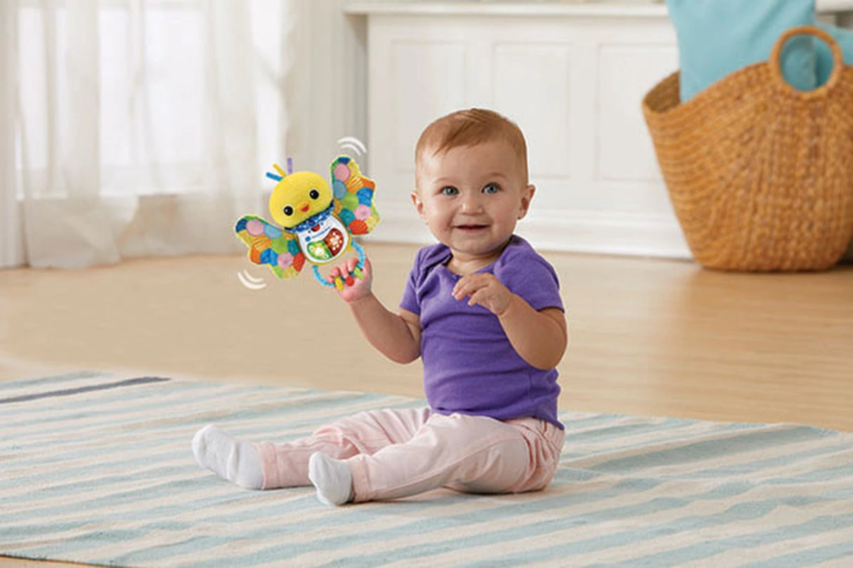 Baby sitting on striped rug holding VTech Soft Singing Birdie Rattle with wings spread and motion lines indicating shaking action