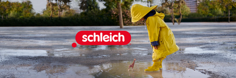 Child in yellow raincoat looking at a small Schleich giraffe figure in a puddle on a wet day in Ireland with Schleich logo visible.