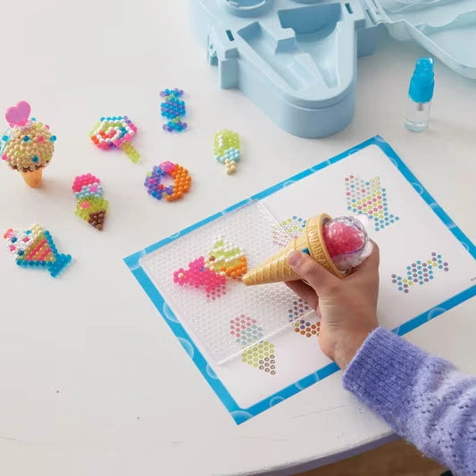 Close-up of a child's hand using the Aquabeads ice cream shaped bead pen to arrange pink and yellow beads on a transparent tray over an illustrated pattern sheet.